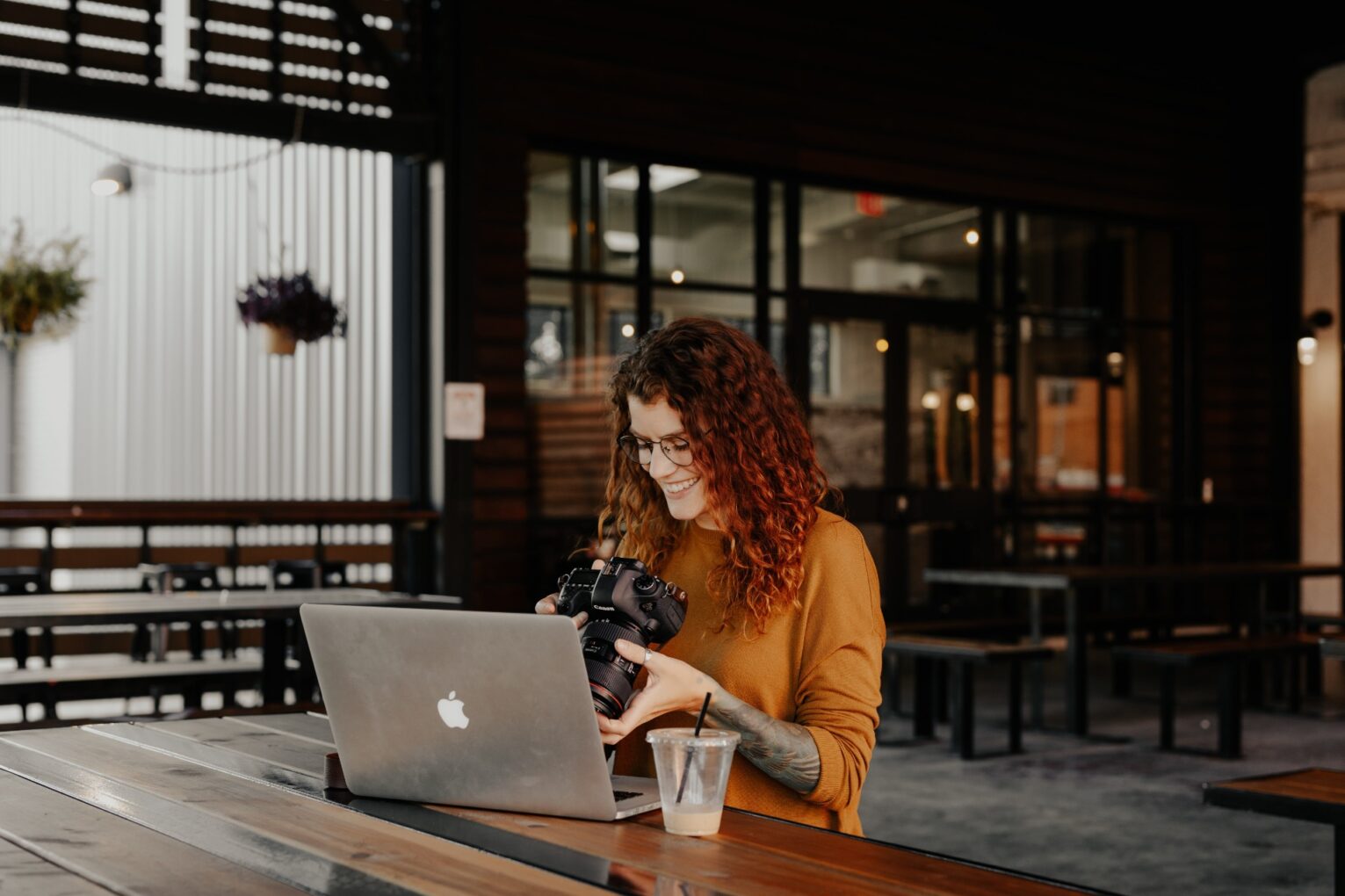 A woman sat in a coffee shop with a MacBook and a camera