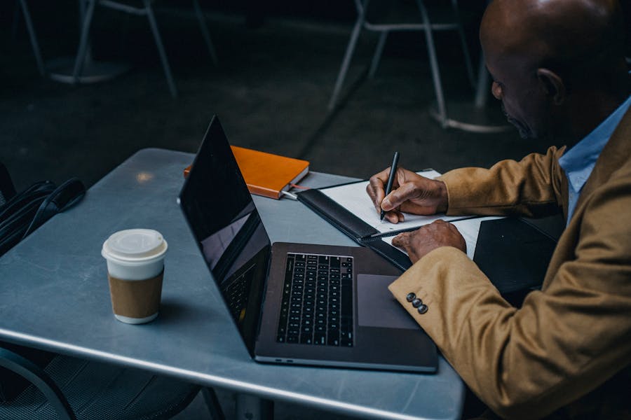 Man researching at table with takeaway coffee cup, laptop and notepad